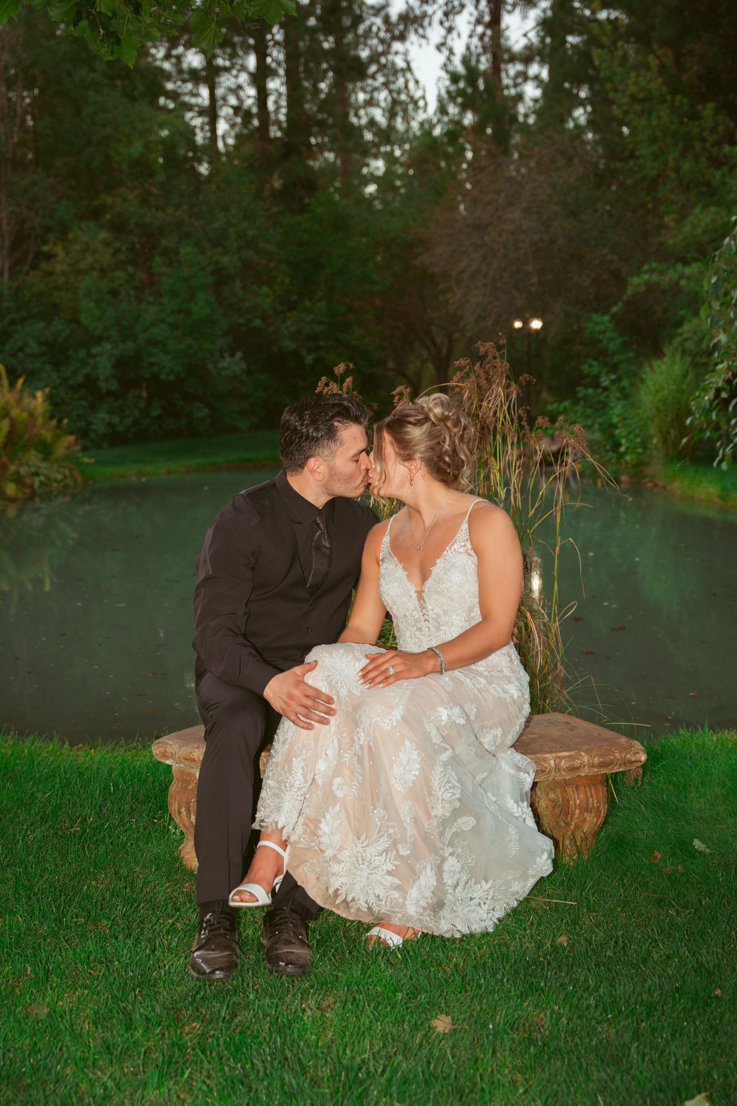 A romantic couple sits together on grass during sunset in a garden setting wearing formal attire.
