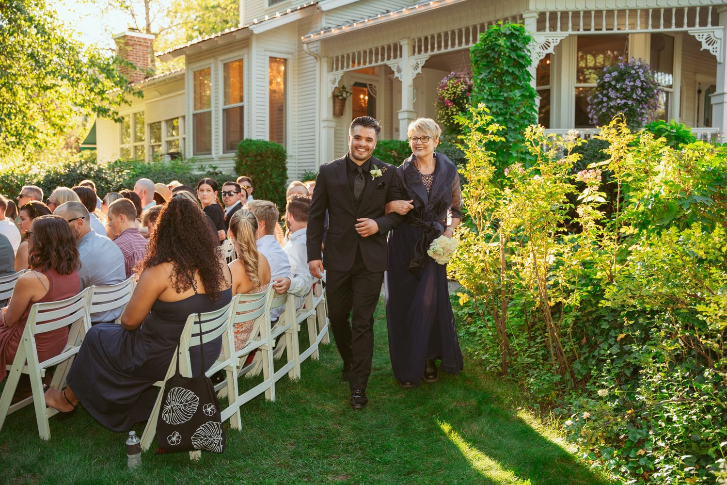Wedding guests gather on a white porch and garden area during an outdoor summer celebration.