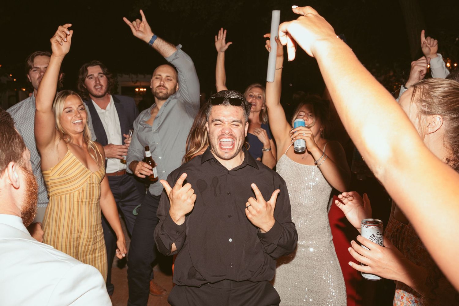 Crowd of wedding guests dancing with raised arms and celebrating at a nighttime outdoor party.