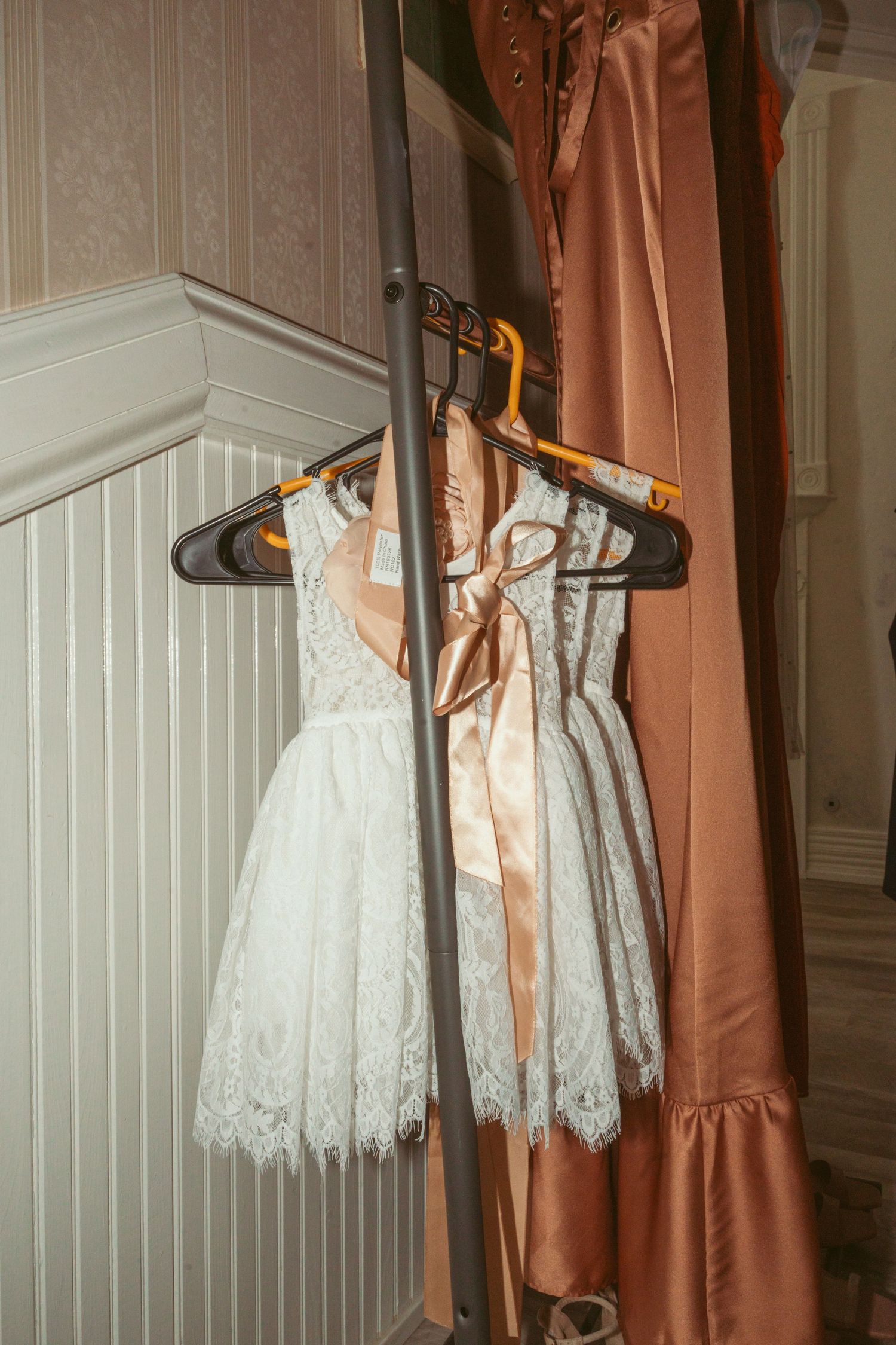 White lace wedding dress and dusty rose bridesmaid dresses hanging on black hangers against a white wall.