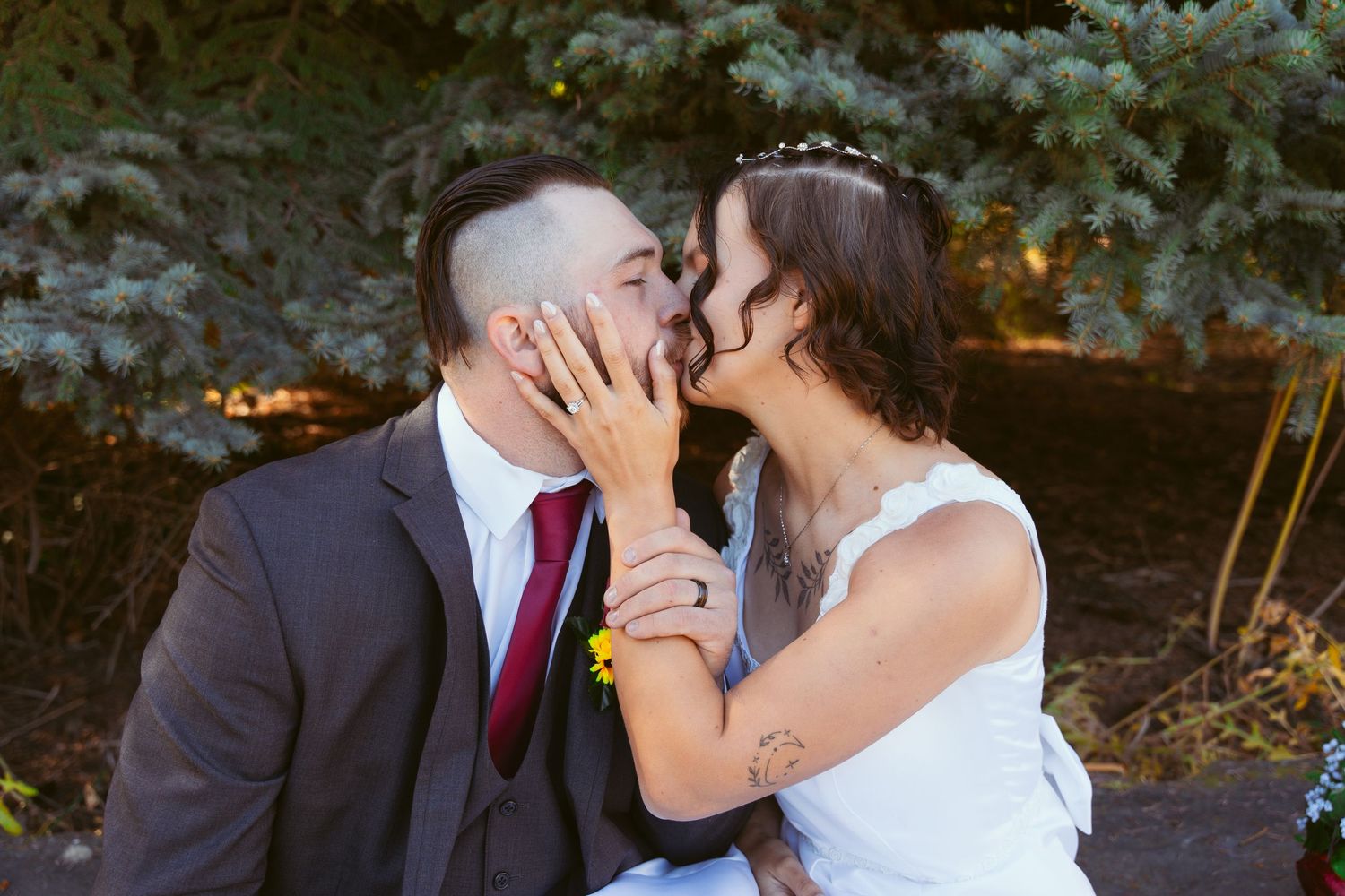 A romantic kiss shared between newlyweds in front of evergreen trees.