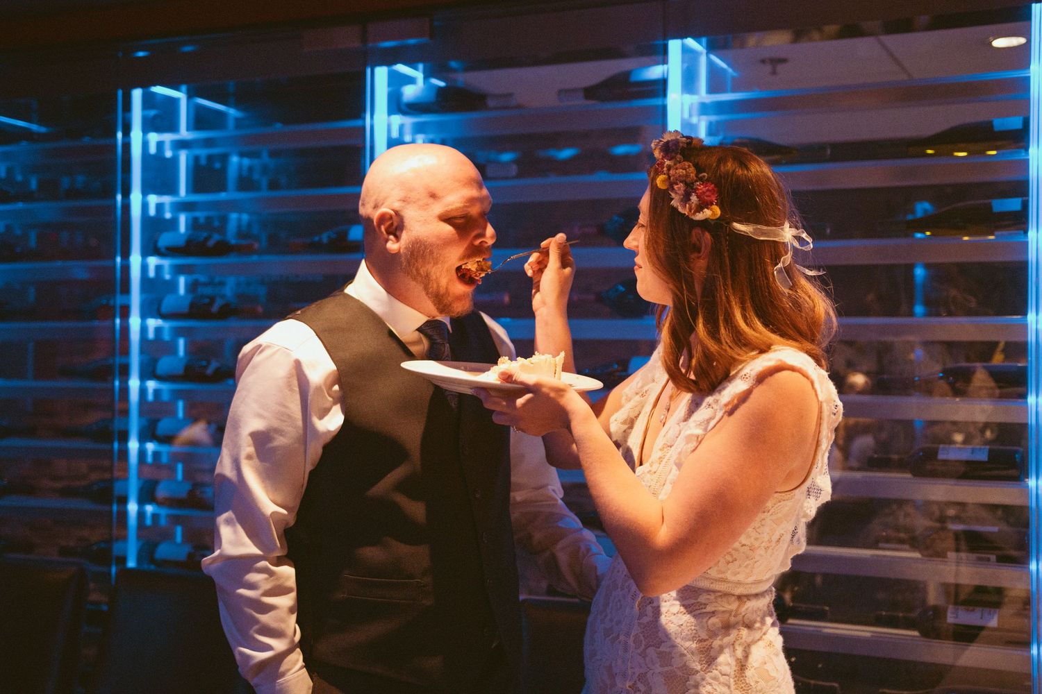 Newlyweds share cake and laughter during their reception under blue lighting.