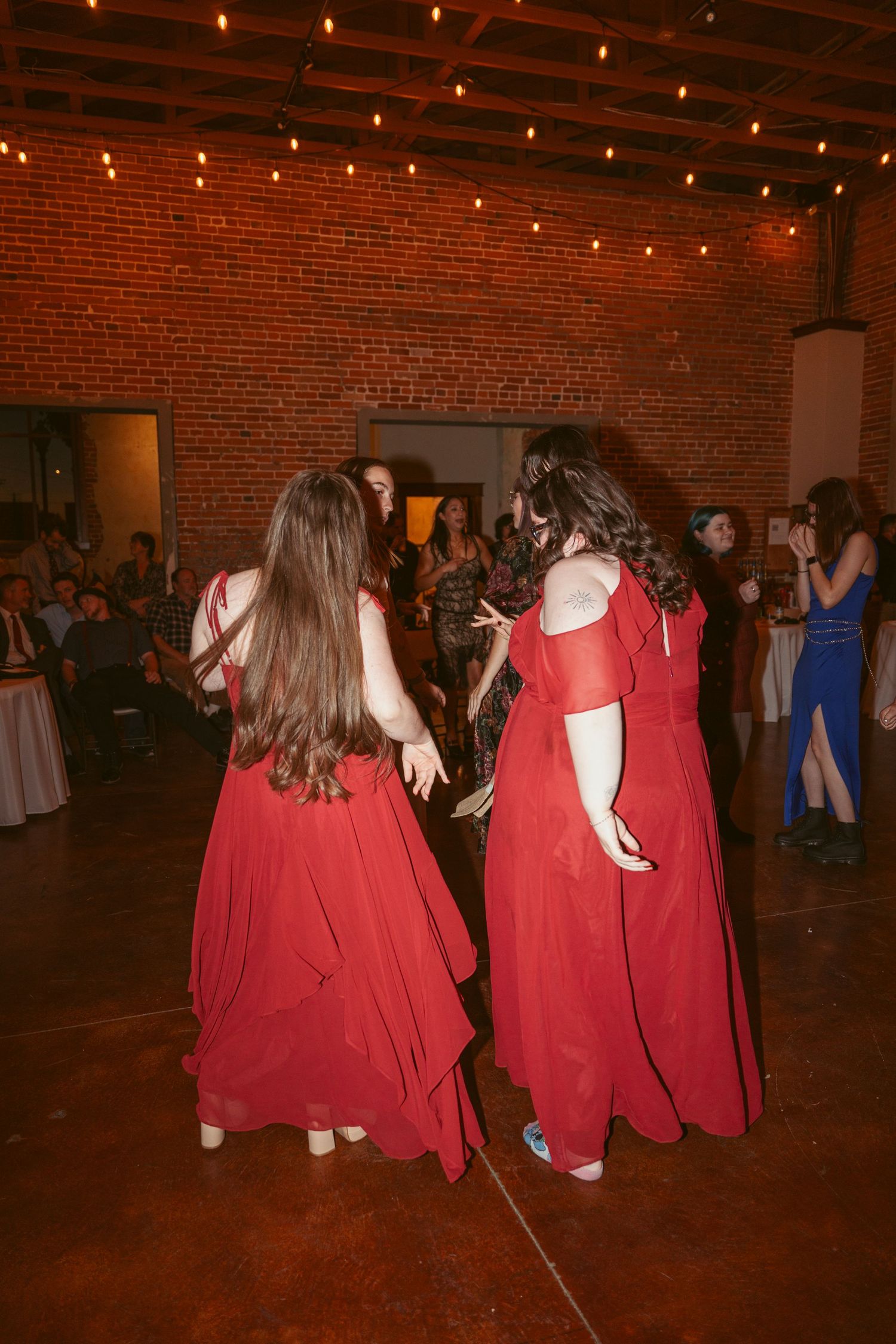 Couples dancing together at an indoor event with exposed brick walls and overhead string lighting.