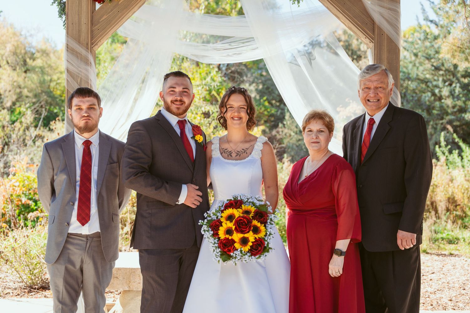 Wedding group photo under rustic wooden pavilion with red and sunflower accents.