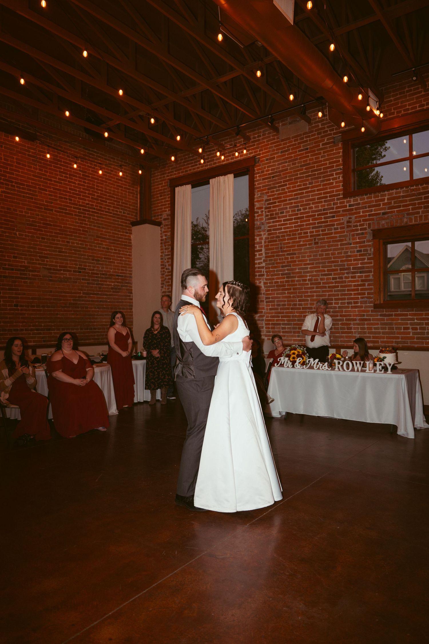 Dance floor moment captured in a rustic reception venue with exposed brick walls and string lights.