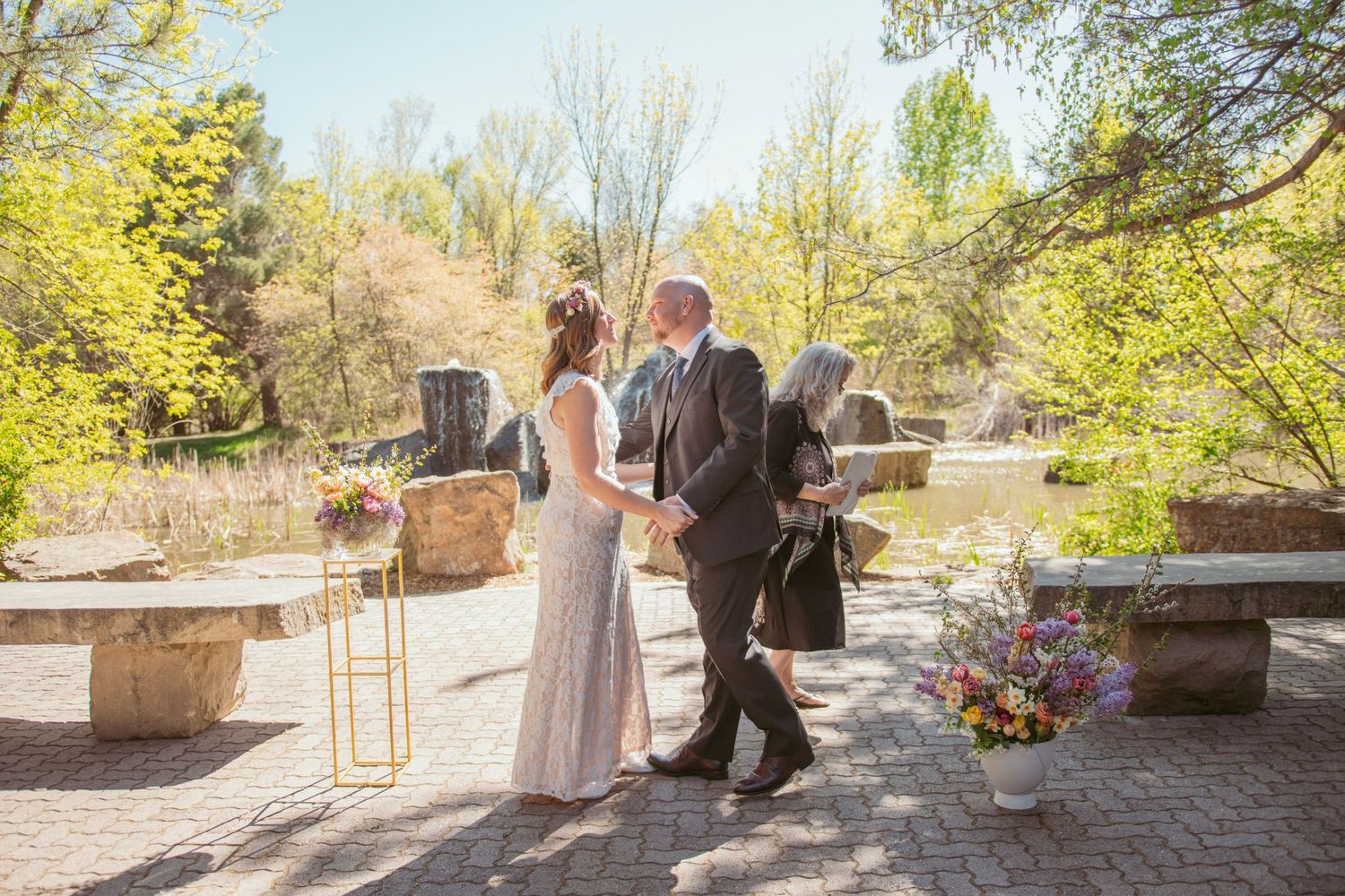 A couple in wedding attire share a romantic moment on a stone patio surrounded by spring foliage.