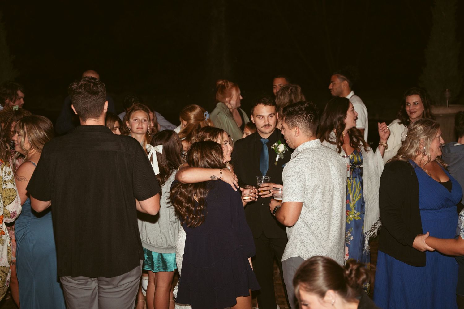 Crowd dancing and celebrating at an outdoor evening event with string lights overhead.
