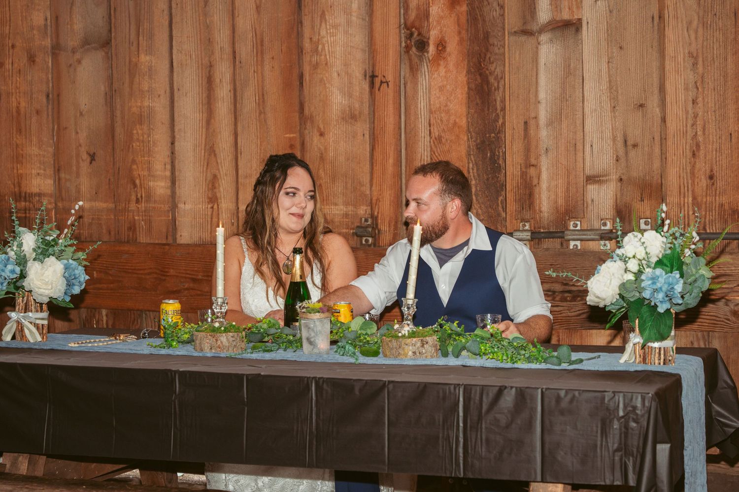 Wedding reception head table decorated with floral arrangements in a rustic barn setting.