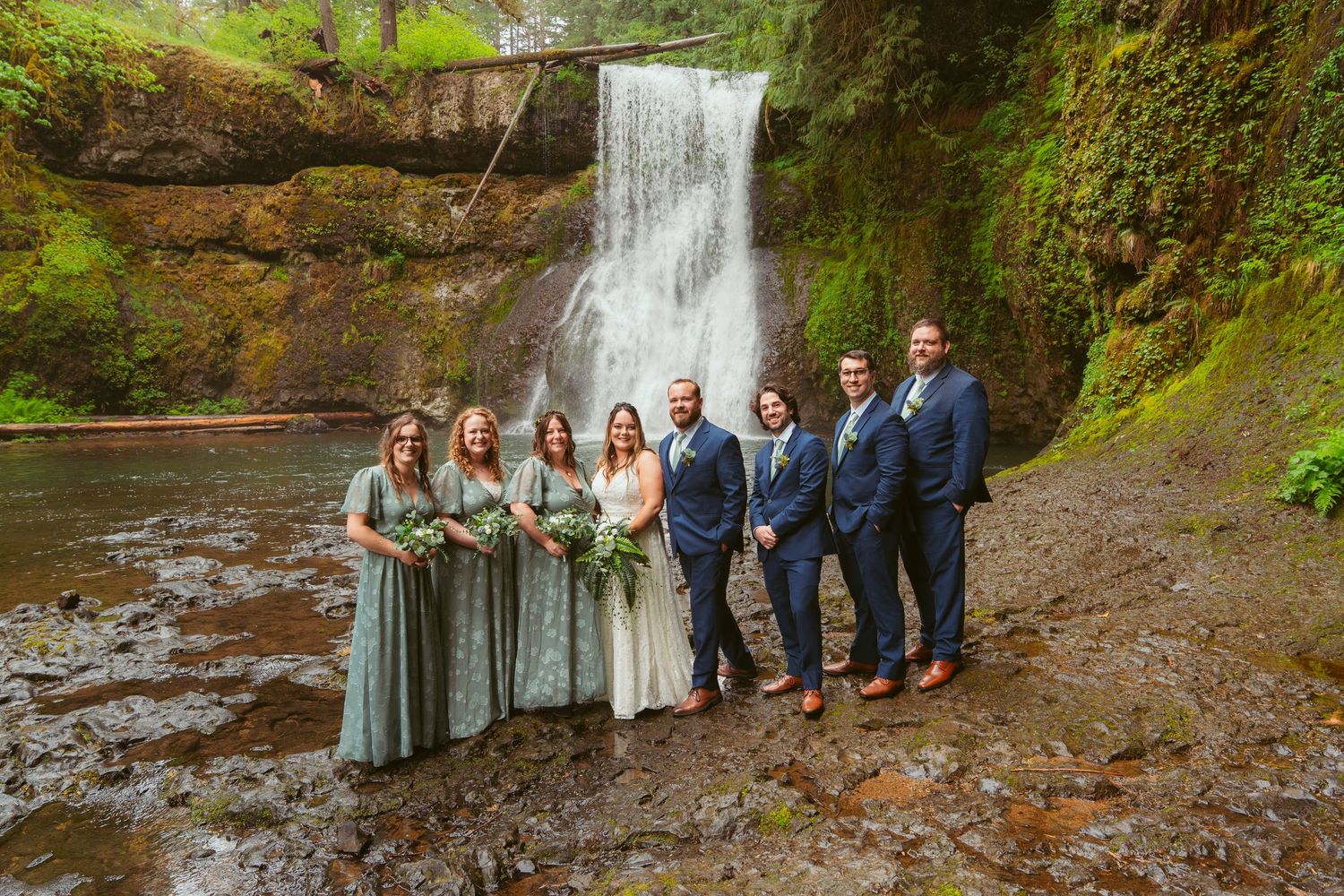 Wedding party poses in front of a scenic waterfall wearing sage green and navy blue attire.