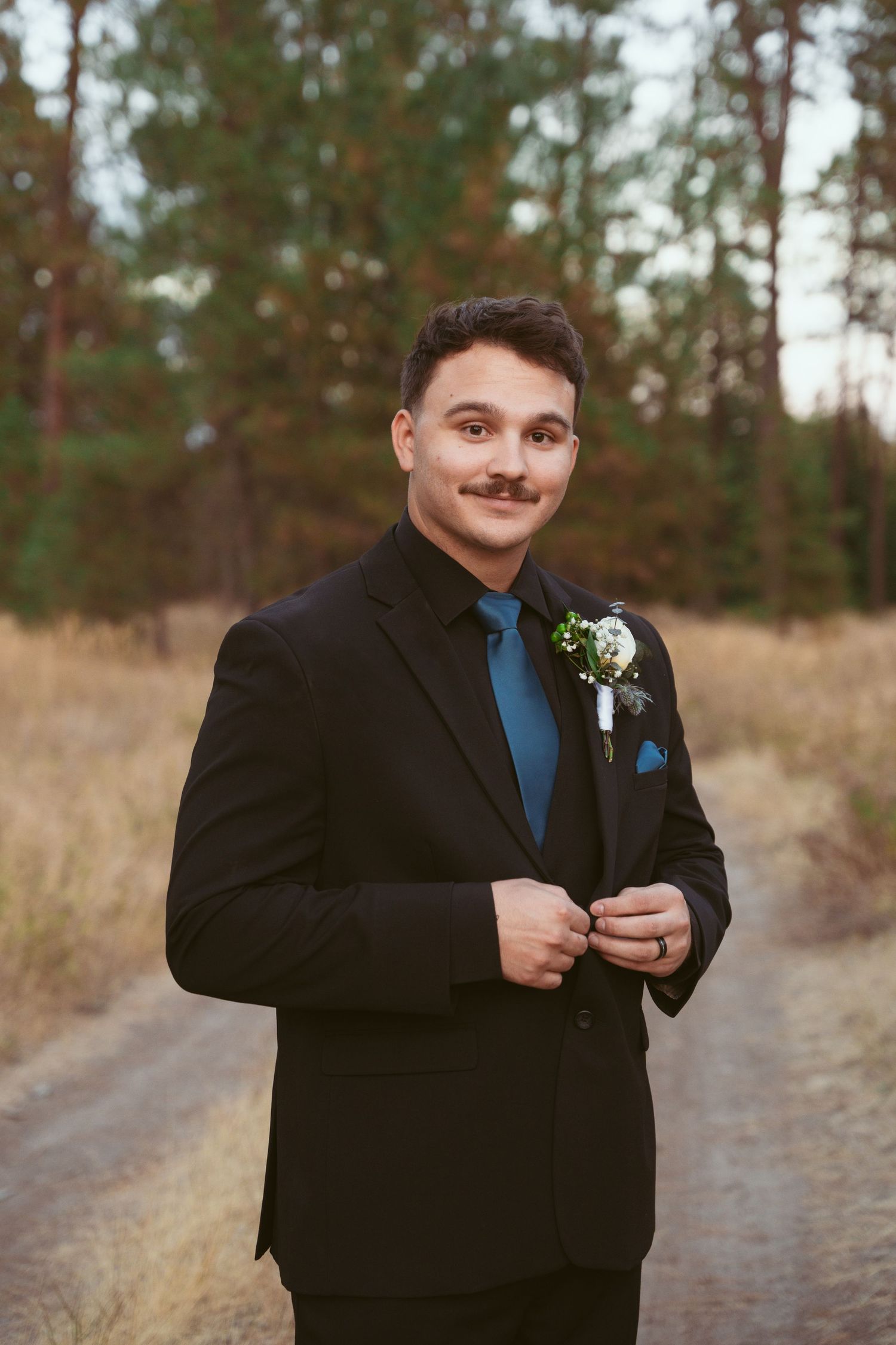 A groom in a black tuxedo and blue tie poses playfully for wedding photos outdoors.