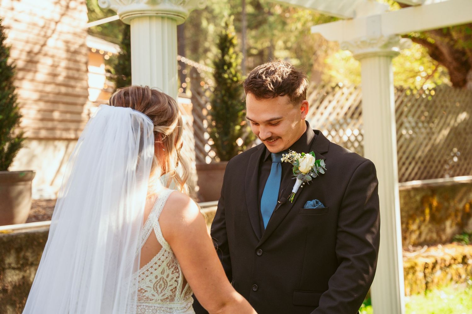 Outdoor wedding ceremony with couple standing beneath white pergola against brick building.