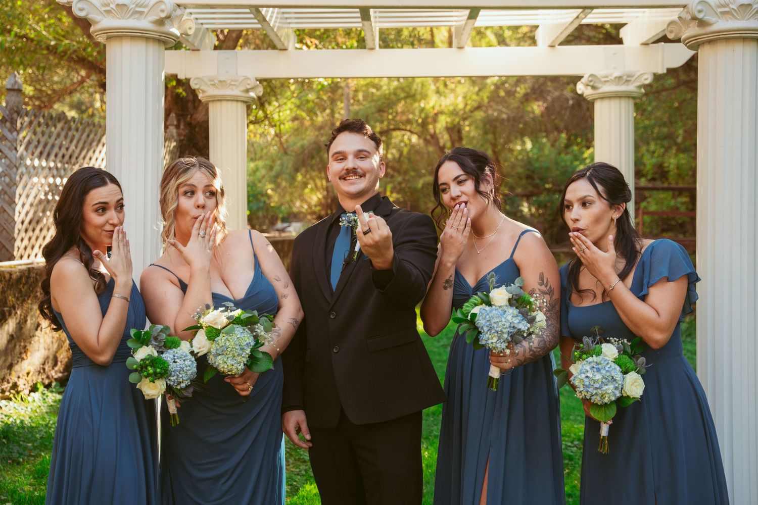 Bridesmaids in matching navy gowns hold blue and white bouquets while standing under pergola.