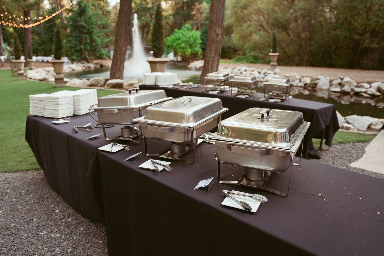 Stainless steel chafing dishes lined up on a dark tablecloth at an outdoor catering buffet setup.
