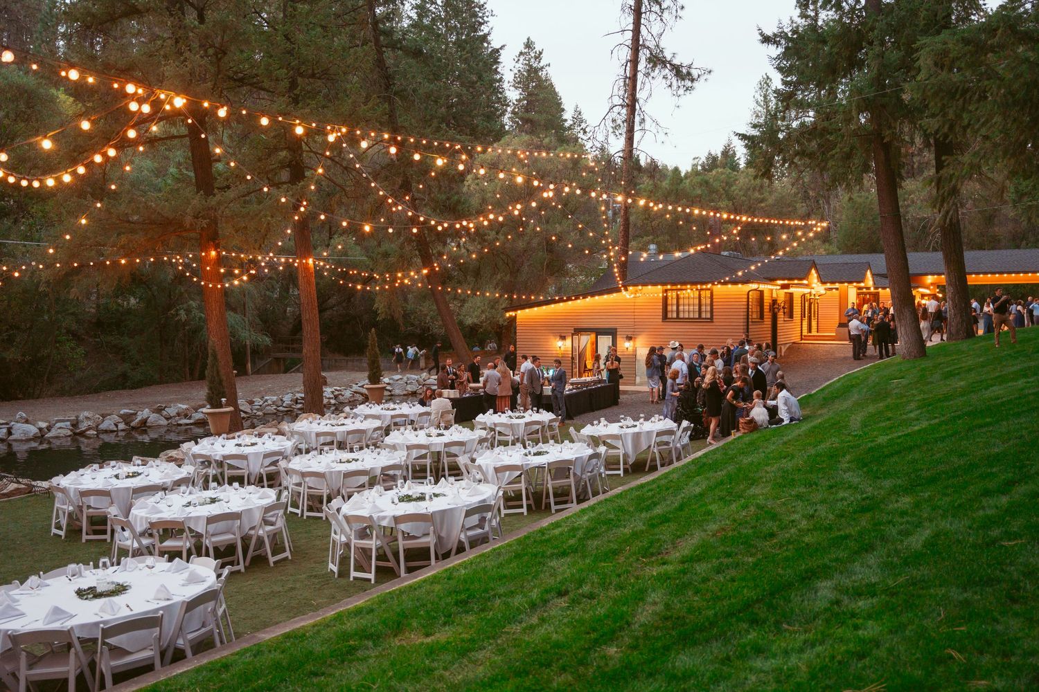 String lights illuminate an outdoor wedding reception venue with white tables and chairs arranged on a grassy hillside.