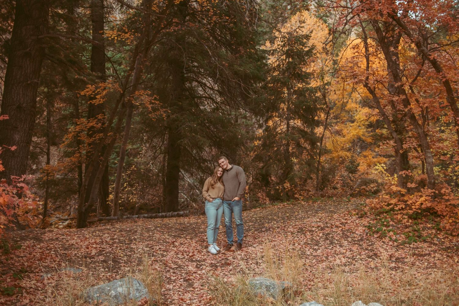 A couple walks through a forest path covered in fallen autumn leaves.
