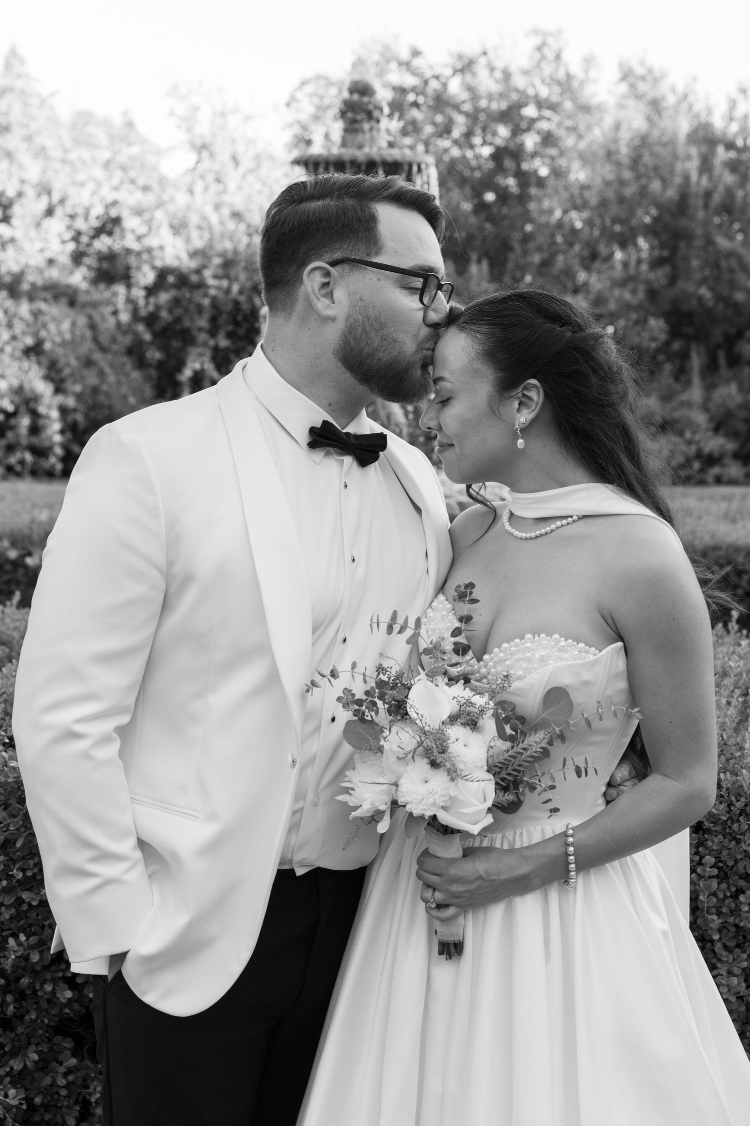 A romantic black and white wedding portrait captured outdoors with a white suit and floral bouquet.