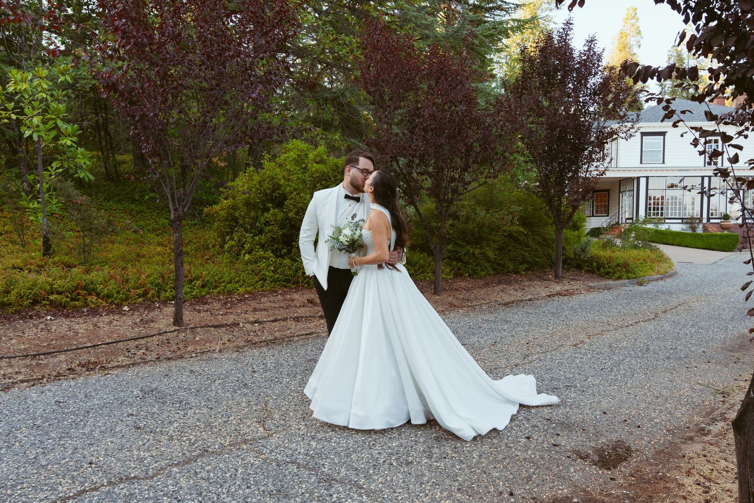A wedding couple walks along a scenic gravel path surrounded by lush greenery and trees.