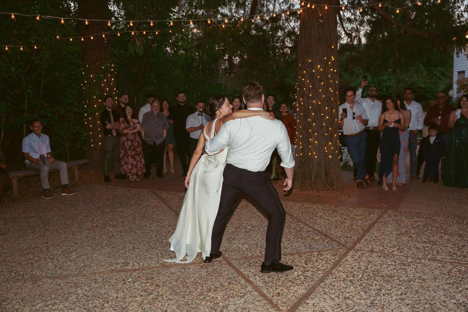 A couple performs their first dance on an outdoor patio under twinkling lights.