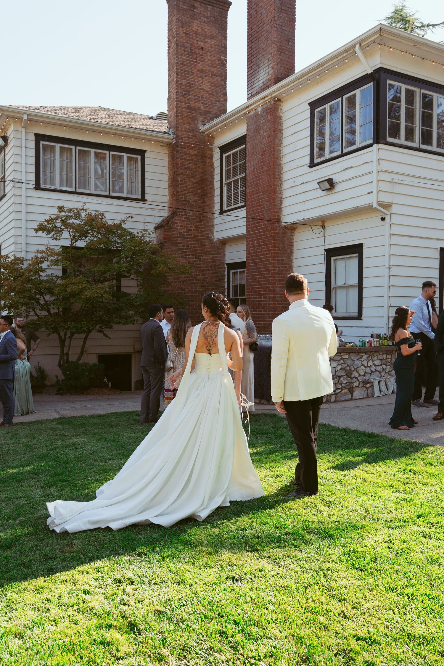A couple in wedding attire walks on a lawn in front of a white building with brick chimneys.