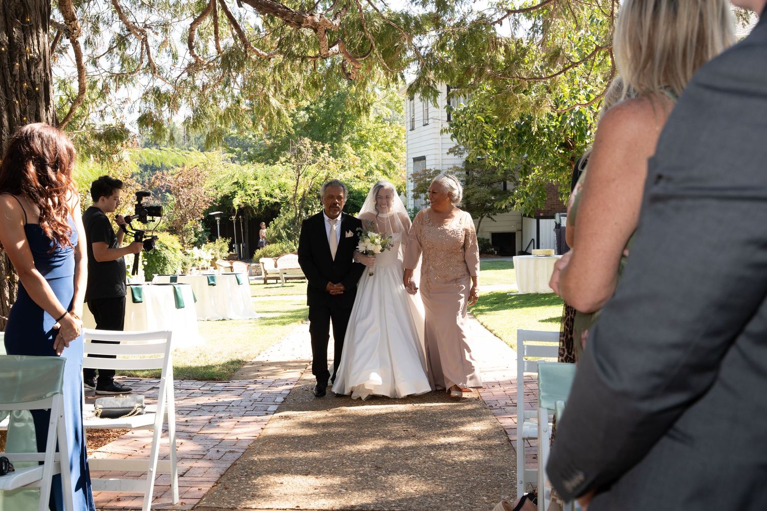 Wedding guests mingle and celebrate at an outdoor garden reception under tall trees with white decorations.