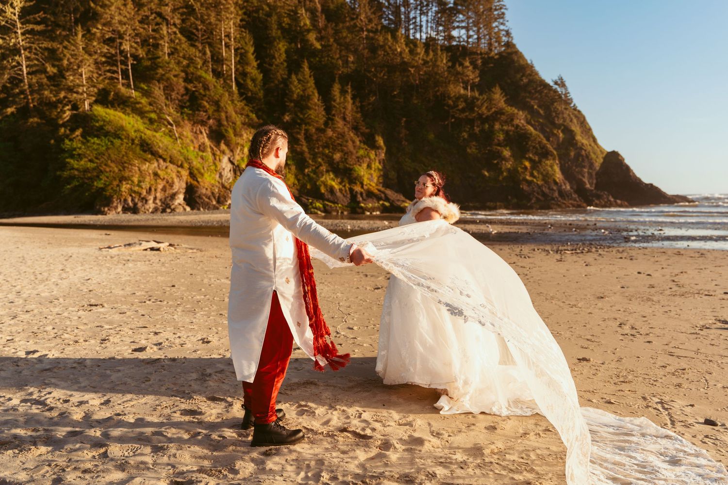 A couple dances with flowing fabric on a scenic beach with mountains and architectural elements in view.