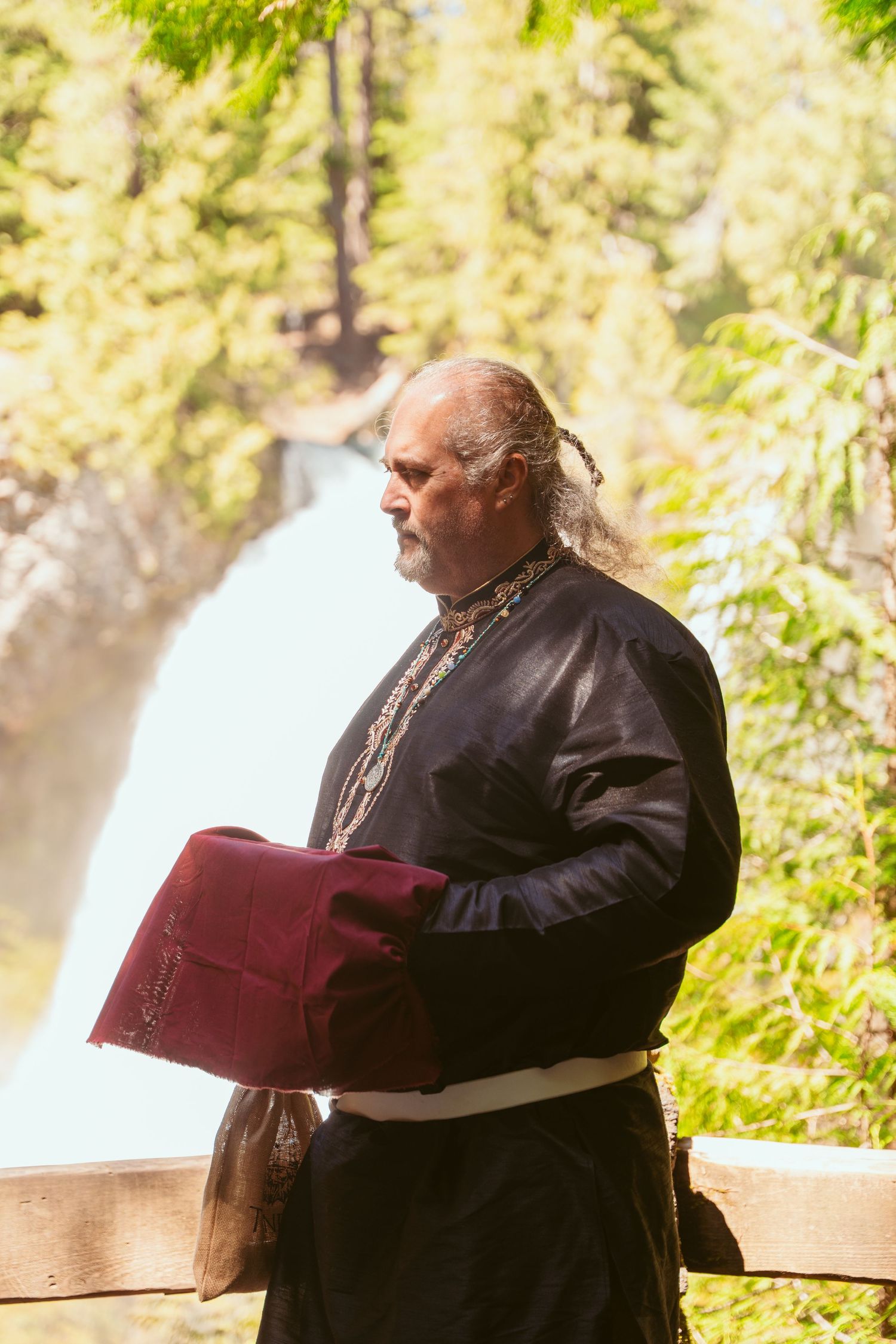 An officiant in dark robes holds a burgundy ceremony book during an outdoor forest wedding.