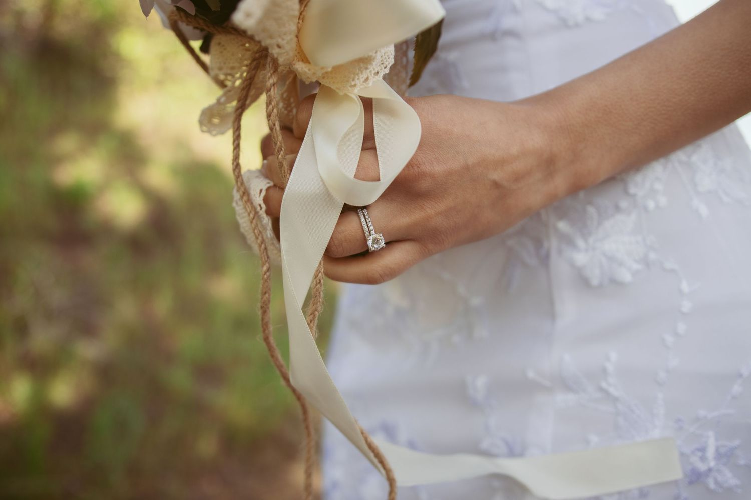 A white bridal bouquet with roses and ribbons held against a natural outdoor background.