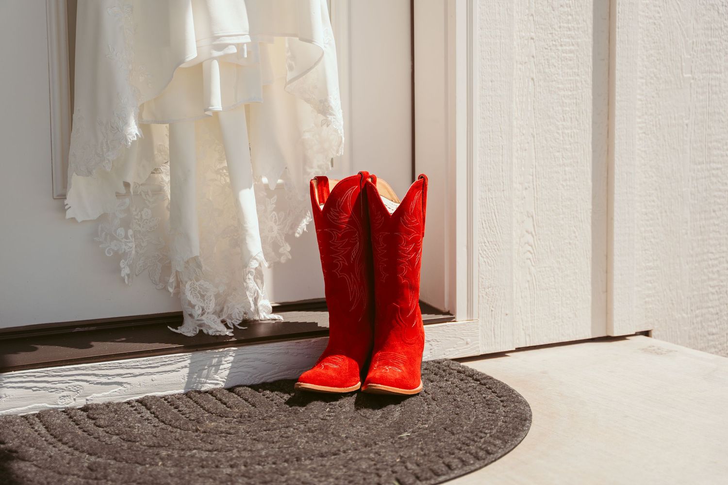 Red cowboy boots next to a white wedding dress on a gray doormat.