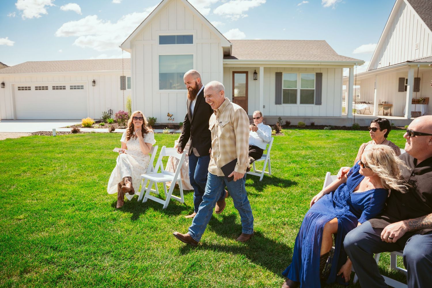 People gathered on the lawn outside a white farmhouse on a sunny day.