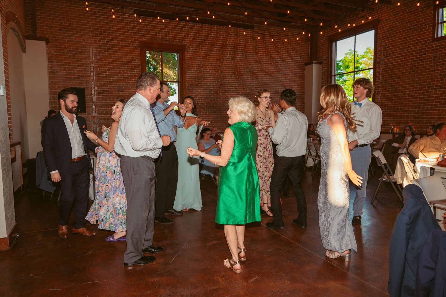 Wedding guests dancing and mingling at an indoor reception with exposed brick walls and string lighting.