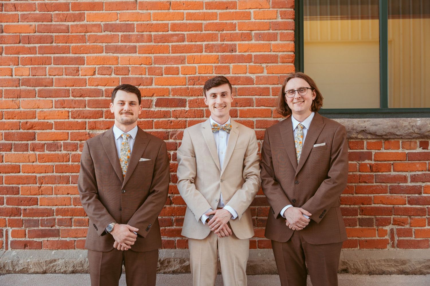 Groomsmen in coordinating brown and beige suits lined up against brick wall.