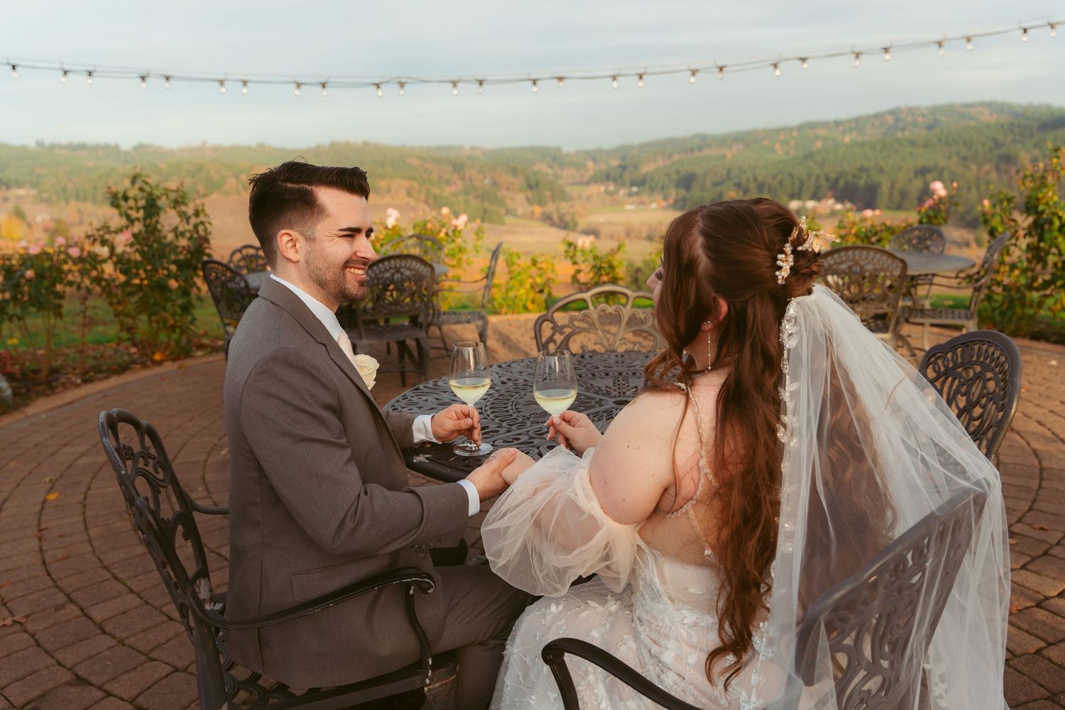 Wedding couple poses for romantic photos on stone patio with string lights and mountain backdrop.