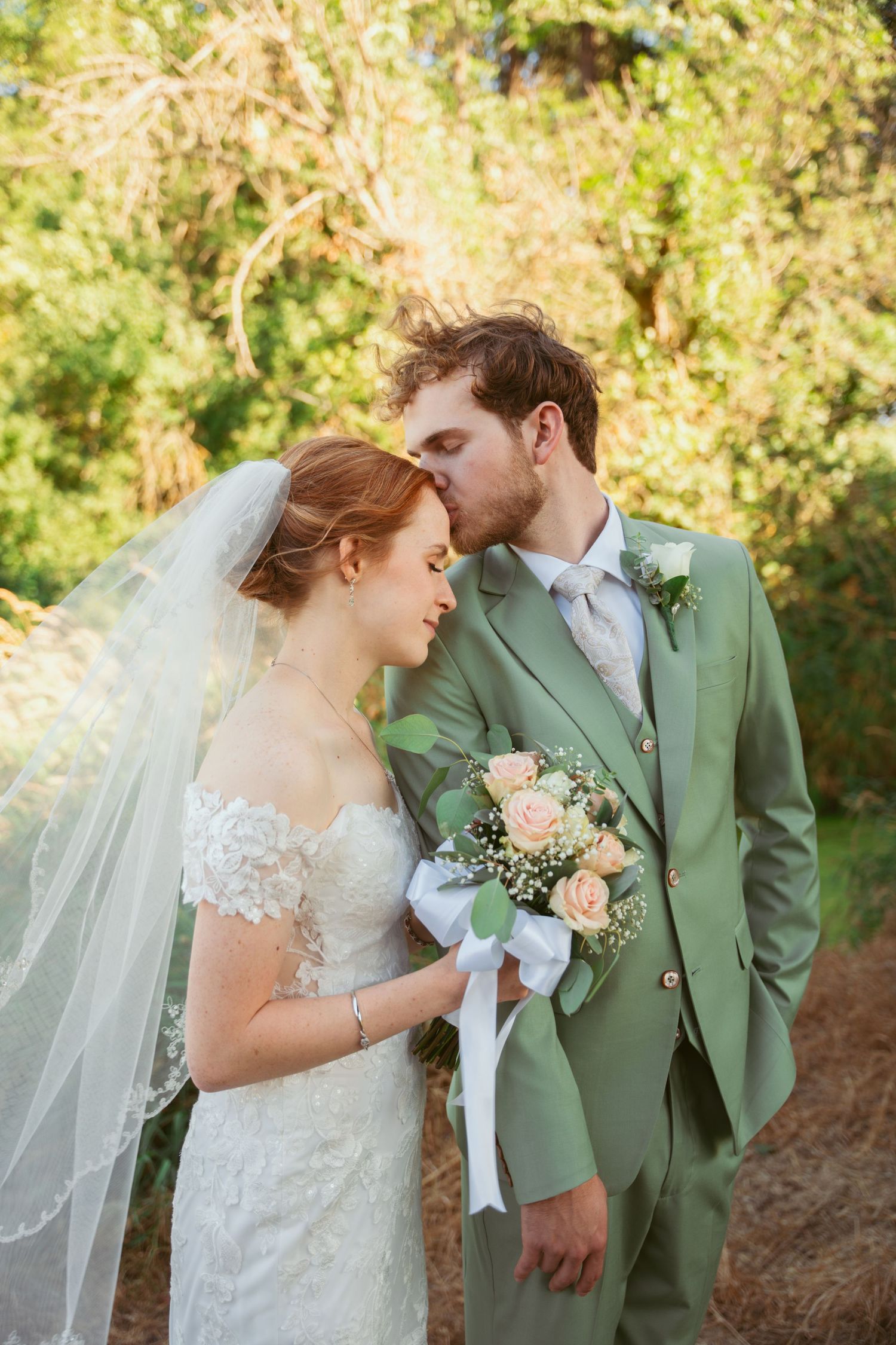 Wedding couple embraces in sage green suit and white lace dress against natural backdrop.