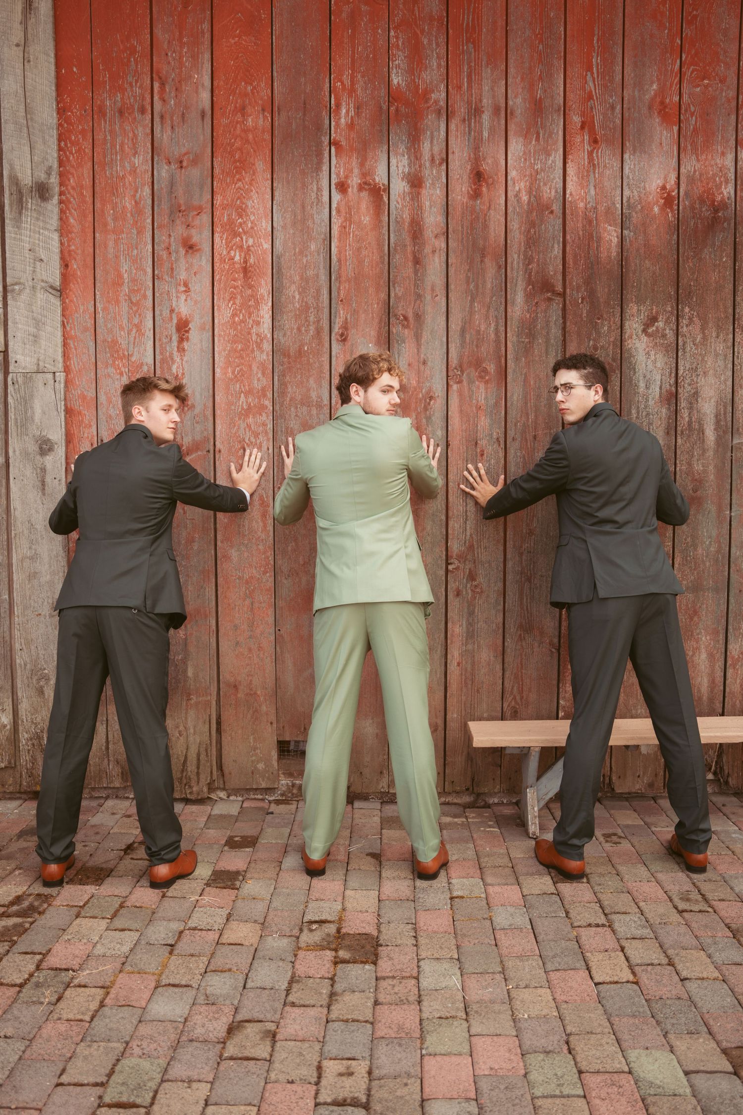 Three men in suits pose humorously against weathered red barn siding.