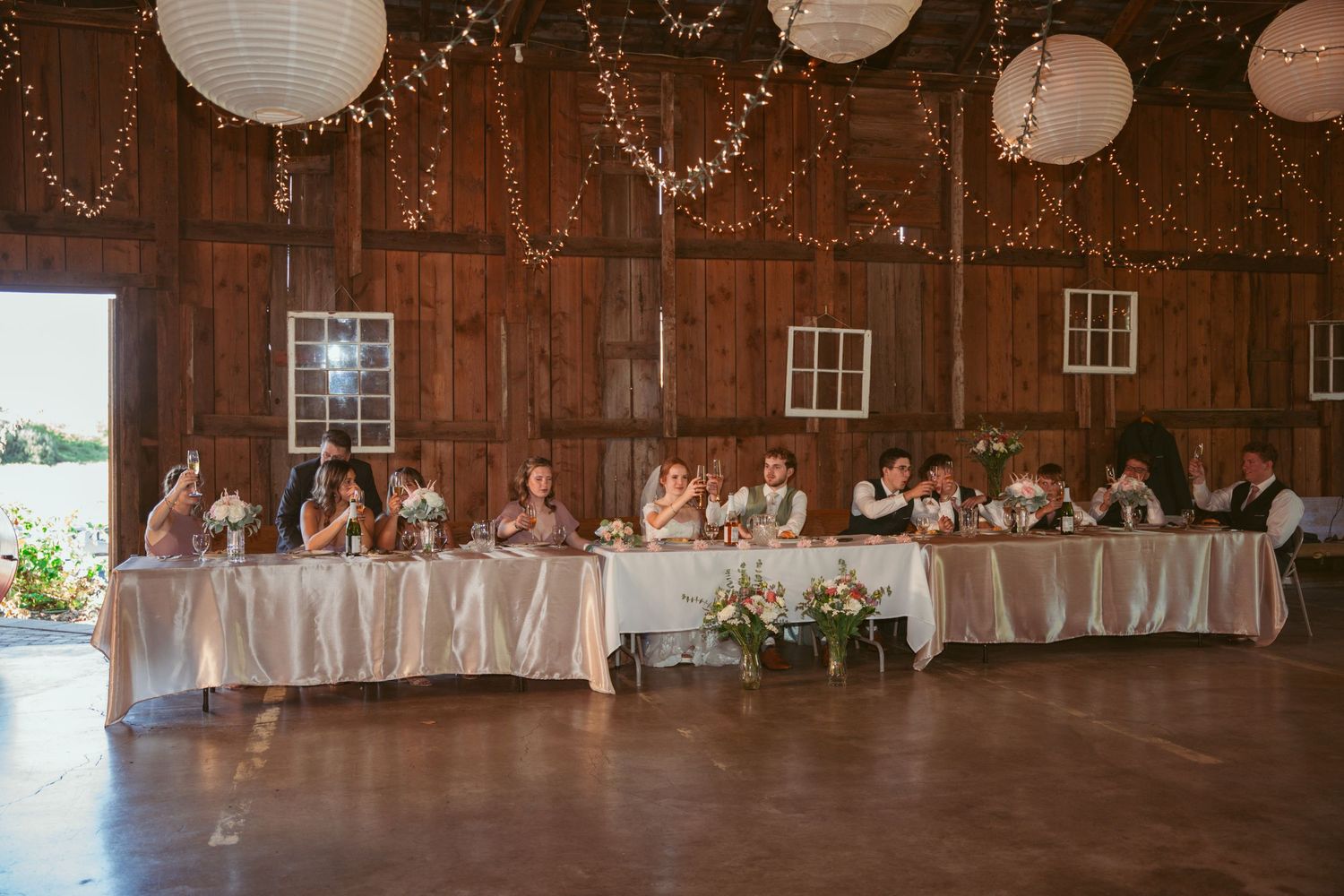 Wedding guests seated at long tables in a rustic barn decorated with twinkling lights.