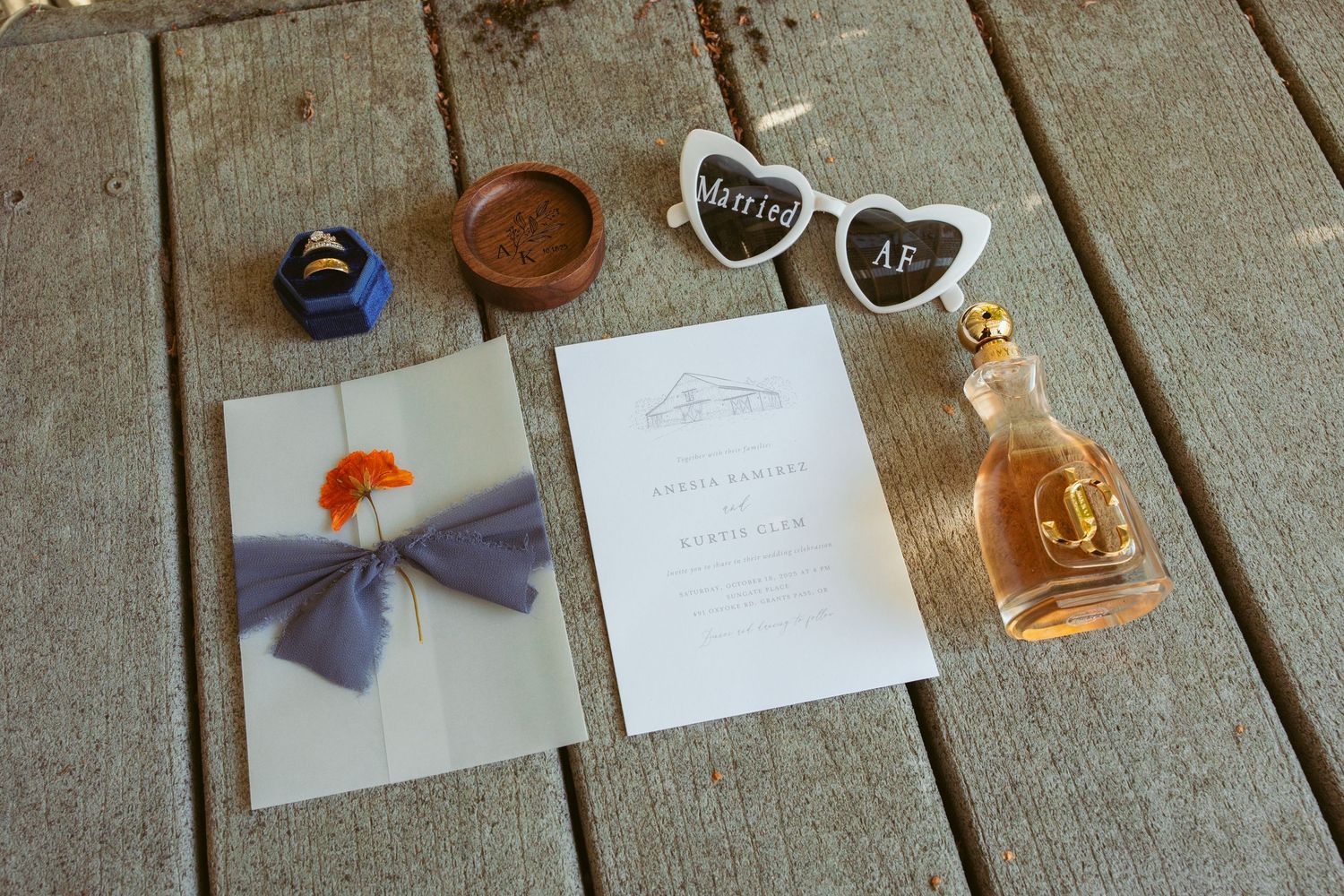 Wedding stationery flat lay with rings, orange flower, and small bottle on wooden deck.