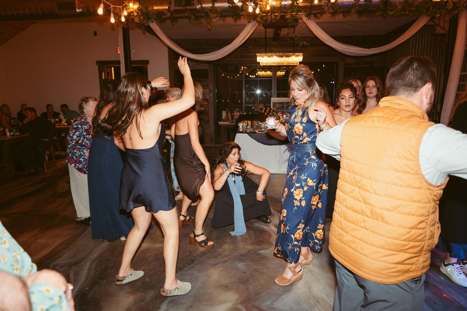Wedding guests dance energetically under string lights and white fabric draping at an indoor reception.