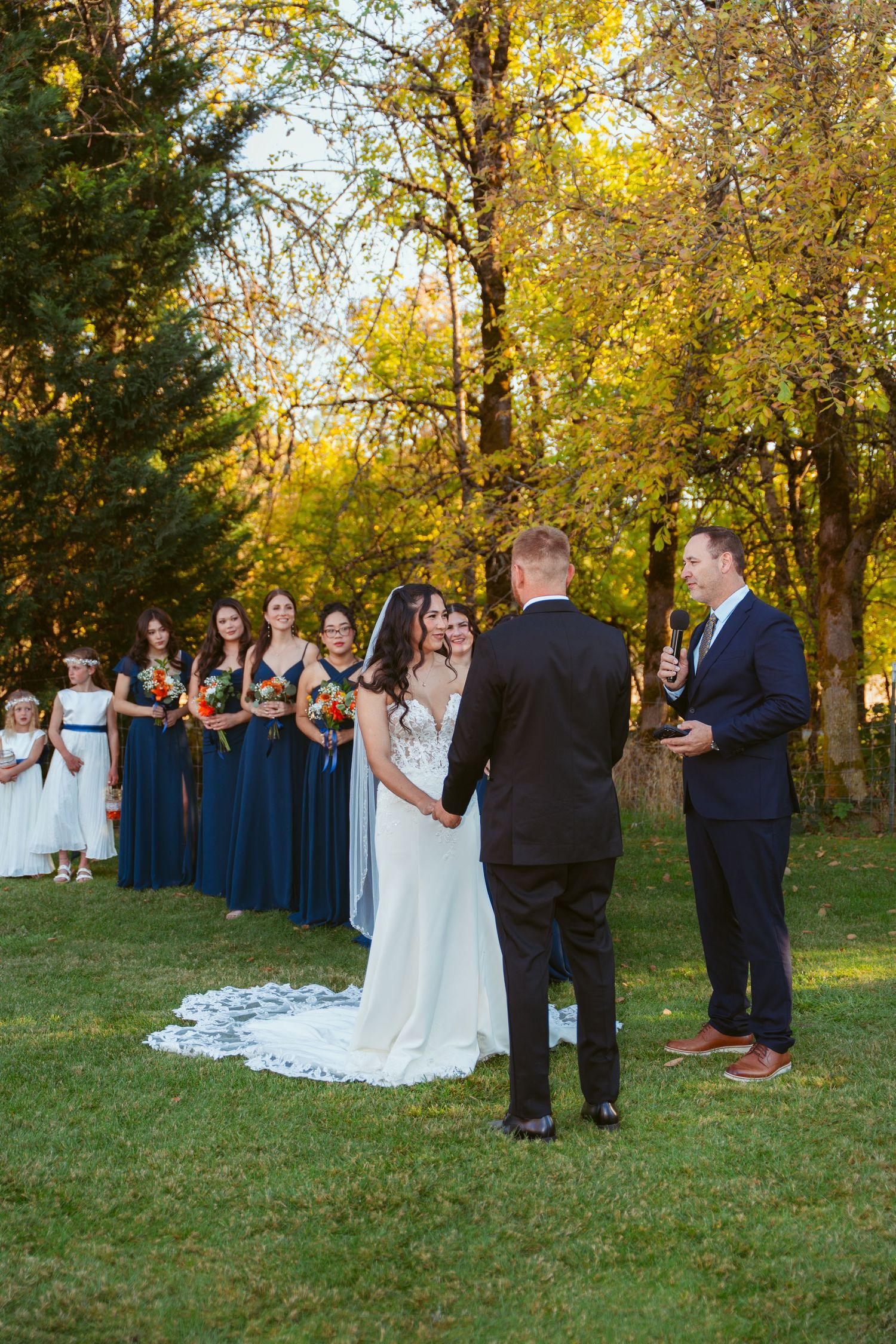 Wedding ceremony in progress with bridal party in navy blue dresses under autumn trees.