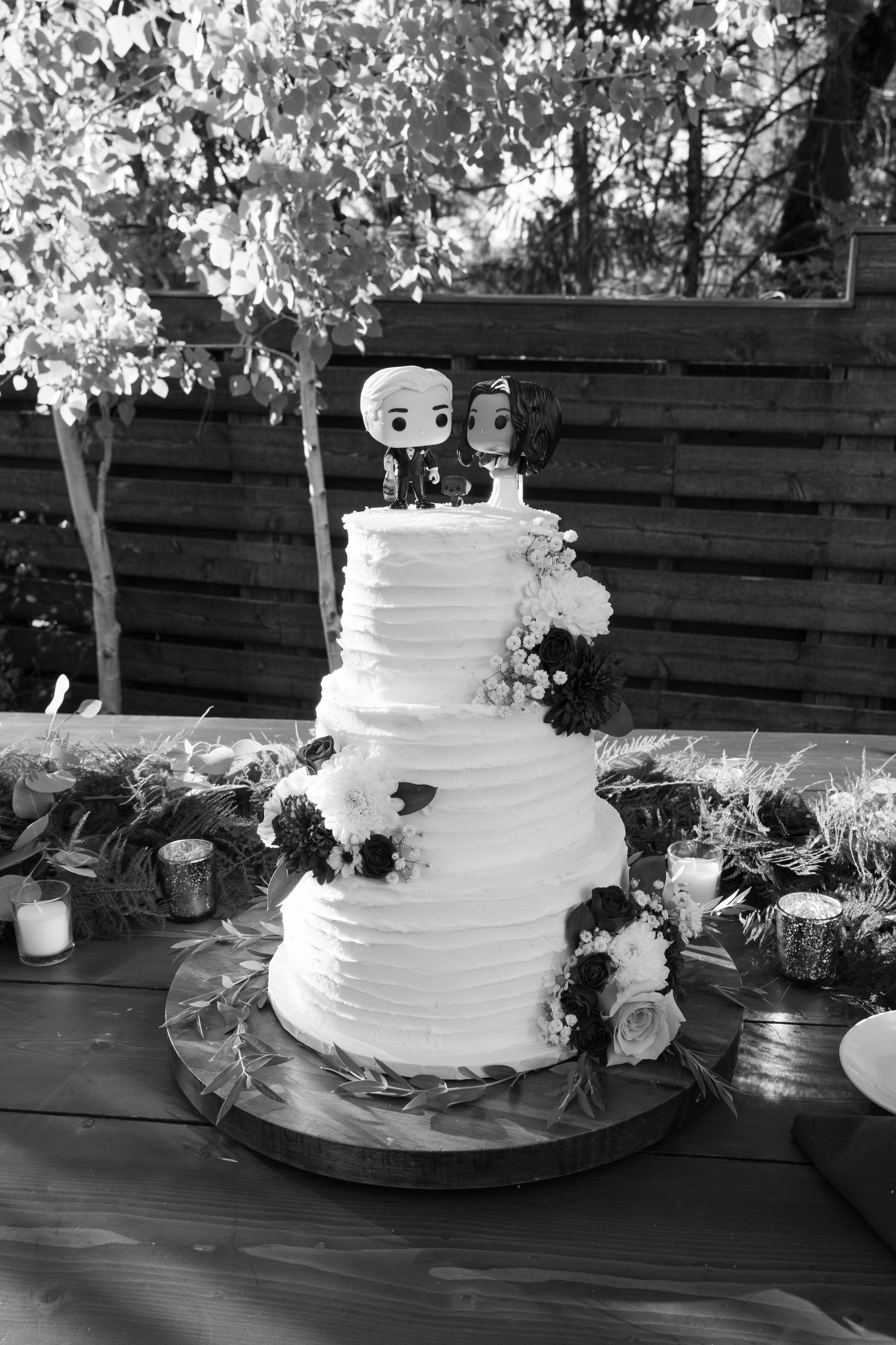 Three-tiered white wedding cake with cartoon cake toppers and floral decorations.