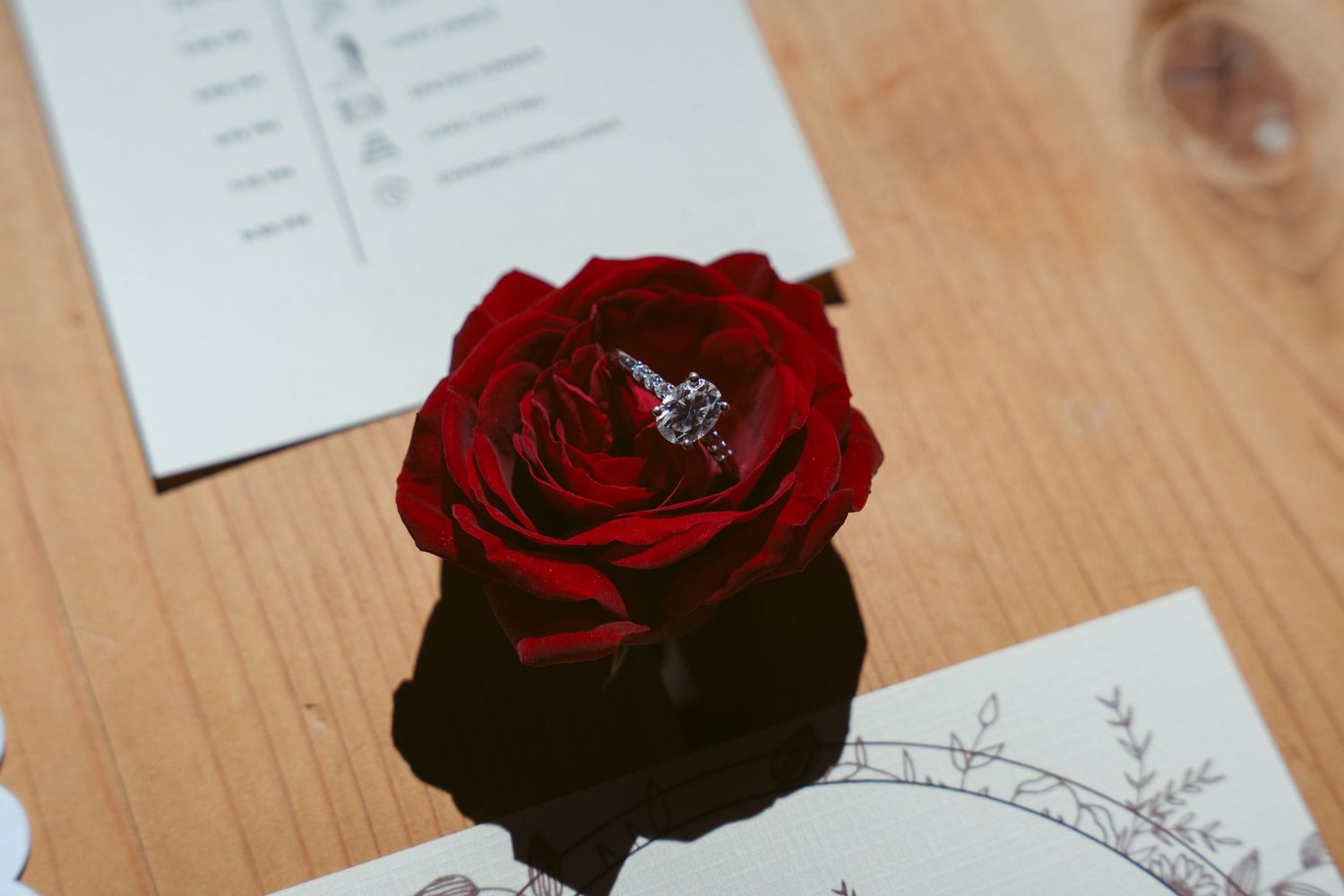 Deep red rose placed next to elegant wedding stationery on wooden surface.