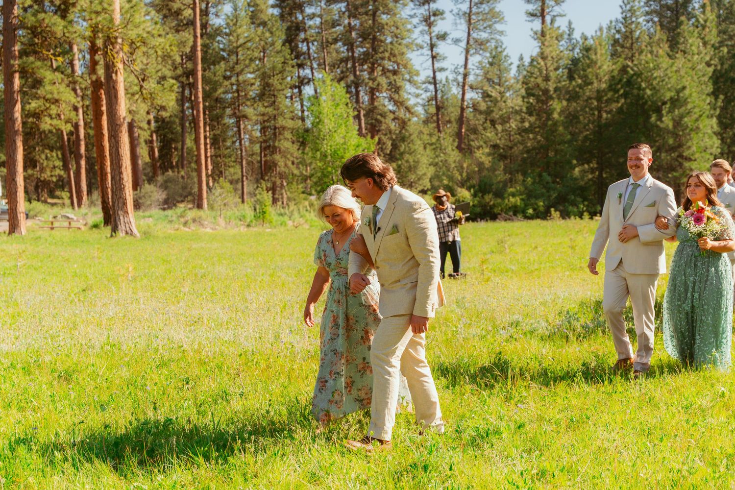 Wedding celebration in a sunny mountain meadow surrounded by tall pine trees.