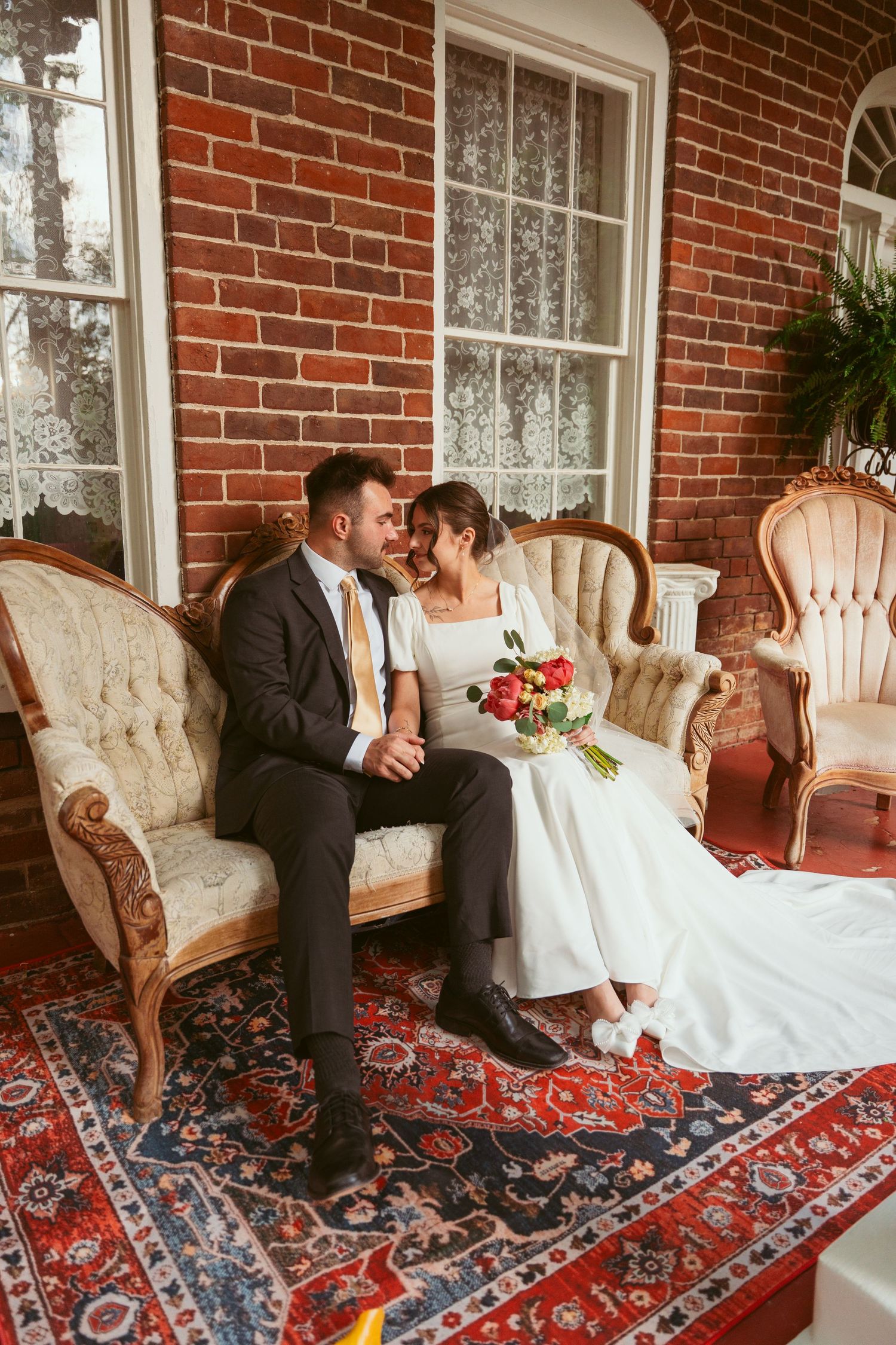 Newlyweds pose together on vintage furniture with elegant Persian rug and brick wall backdrop.