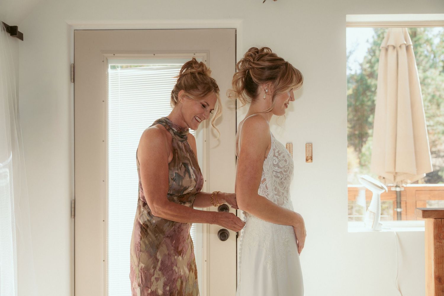 Two people in a sunlit room preparing a white wedding dress.
