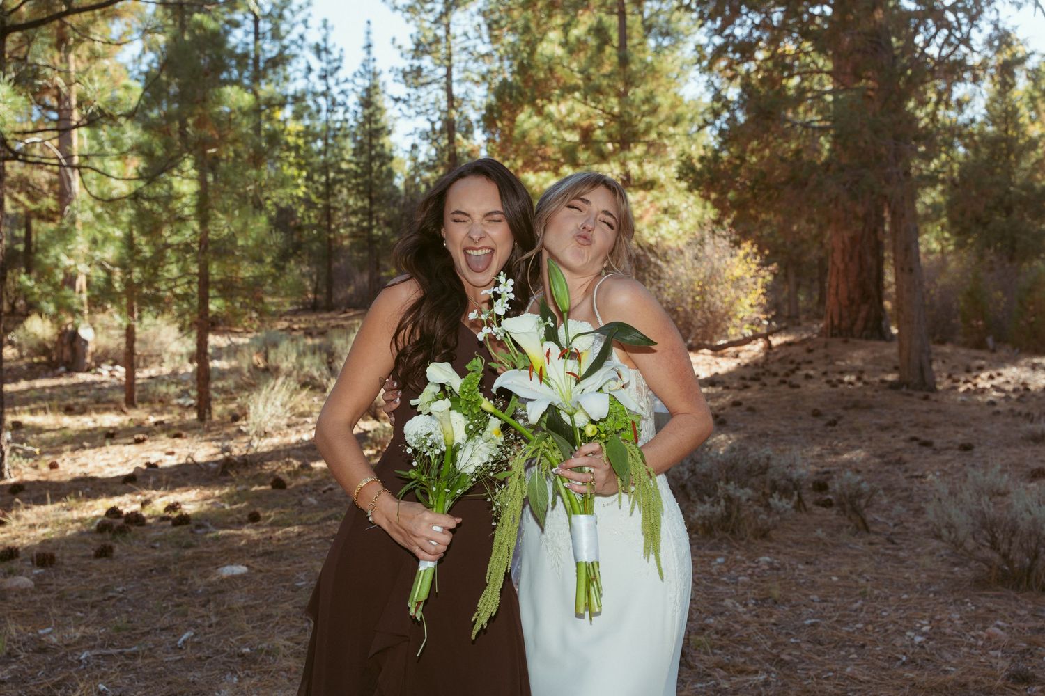 Two people in contrasting outfits share a candid moment while holding white flower bouquets in a forest.