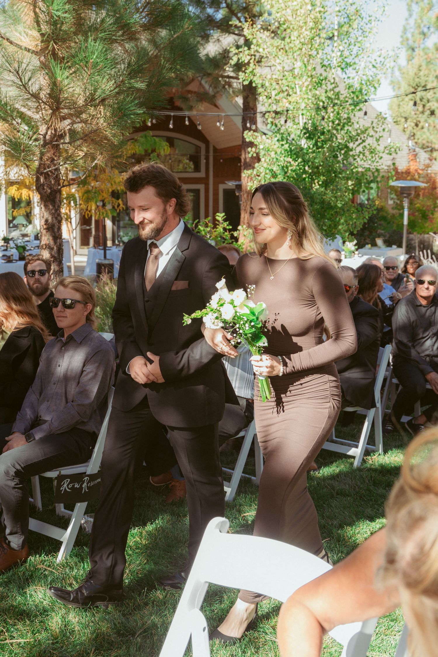 Intimate outdoor wedding ceremony with guests seated on white chairs under leafy trees.