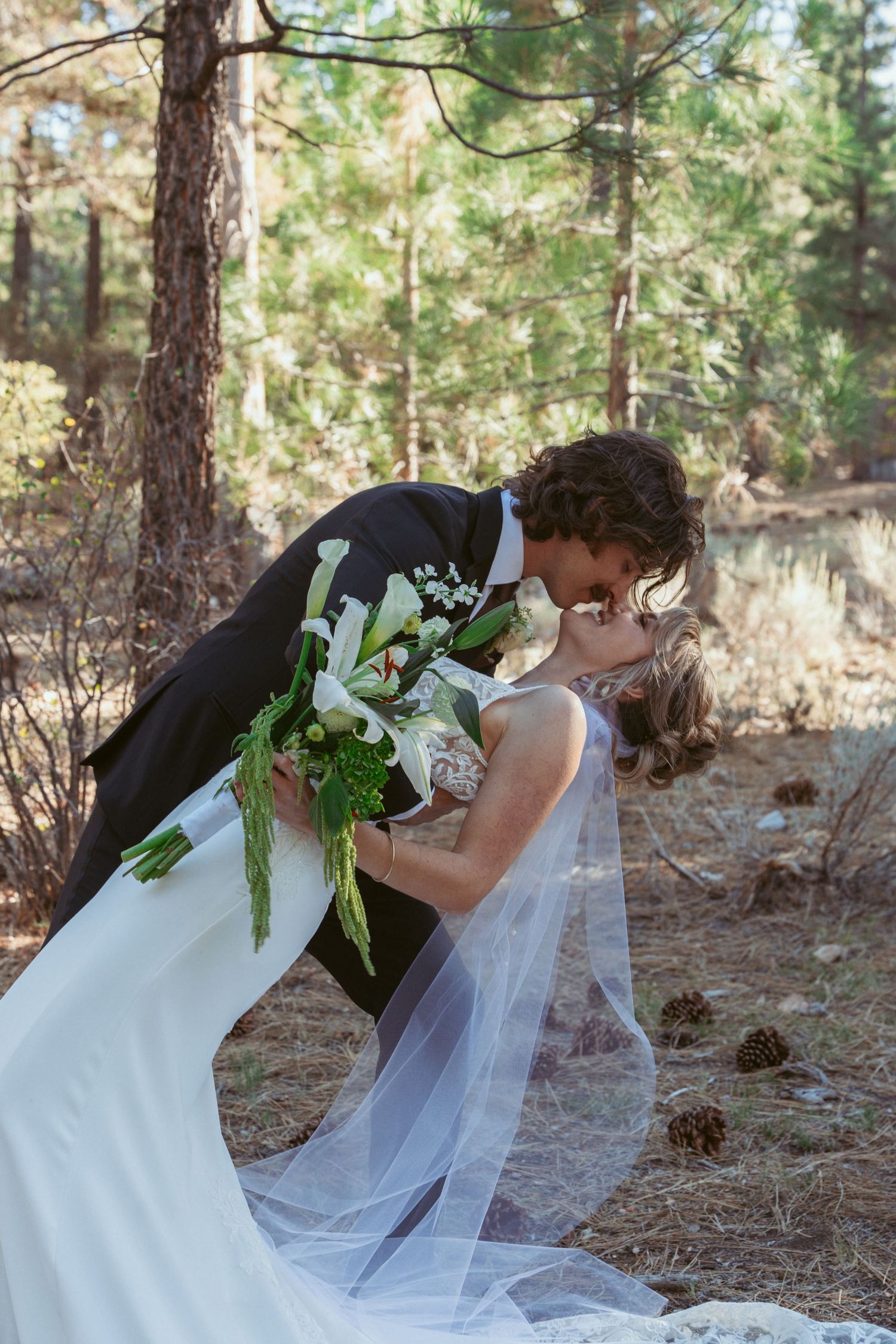 Intimate wedding moment photographed in sunlit forest setting with white veil.