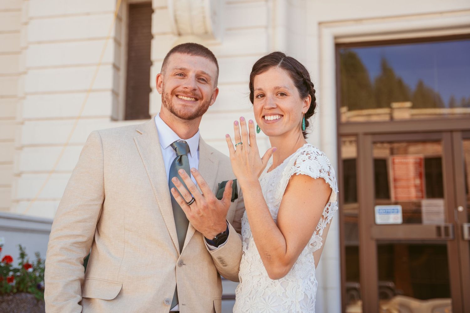 Couple in wedding attire showing off their rings in front of a light colored building.