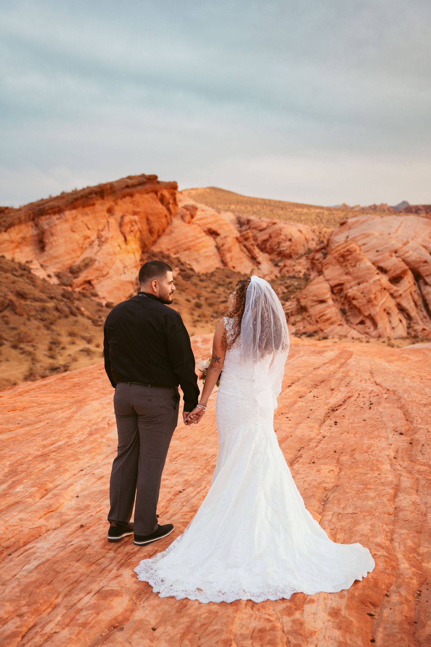 Wedding couple walks hand in hand on red rock formations during sunset.