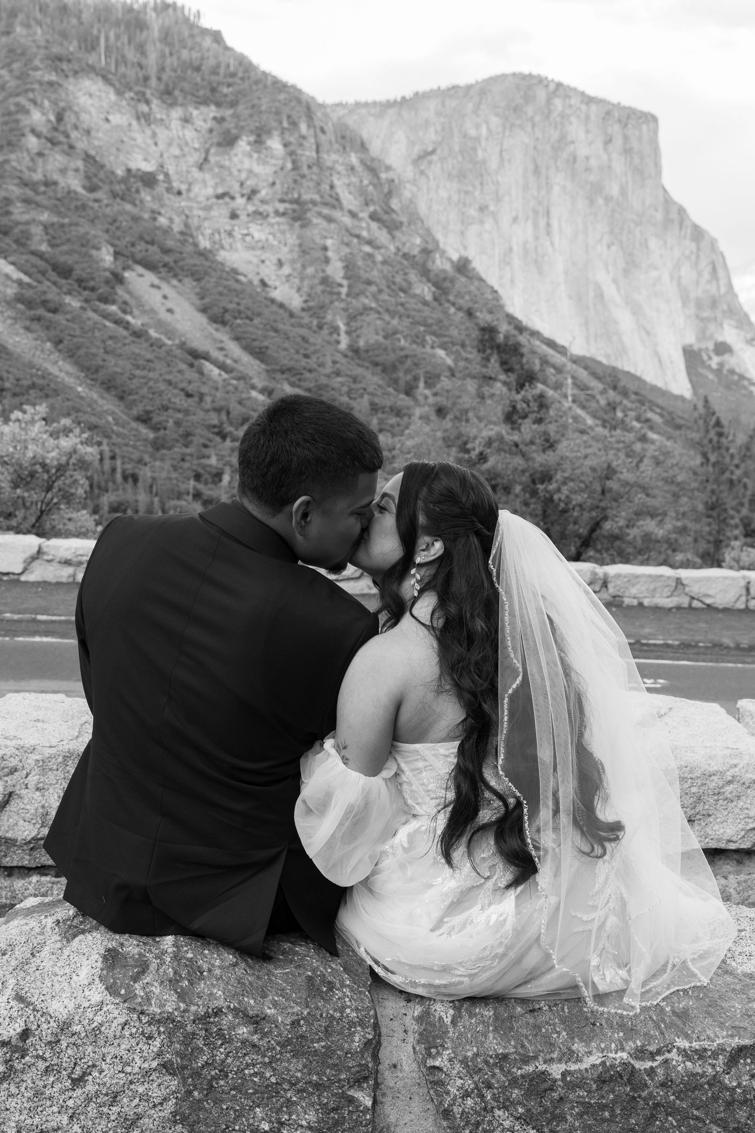 Newlyweds share an intimate moment on a gravel path with towering cliffs in the background.