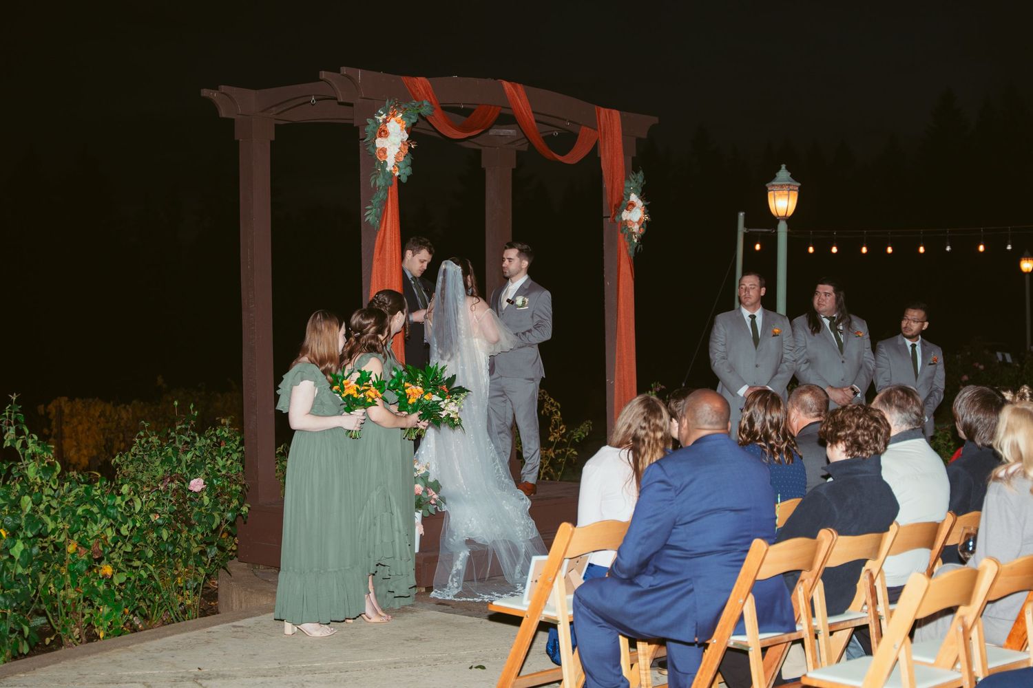 Outdoor wedding venue setup with string lights and red-draped arch at dusk.